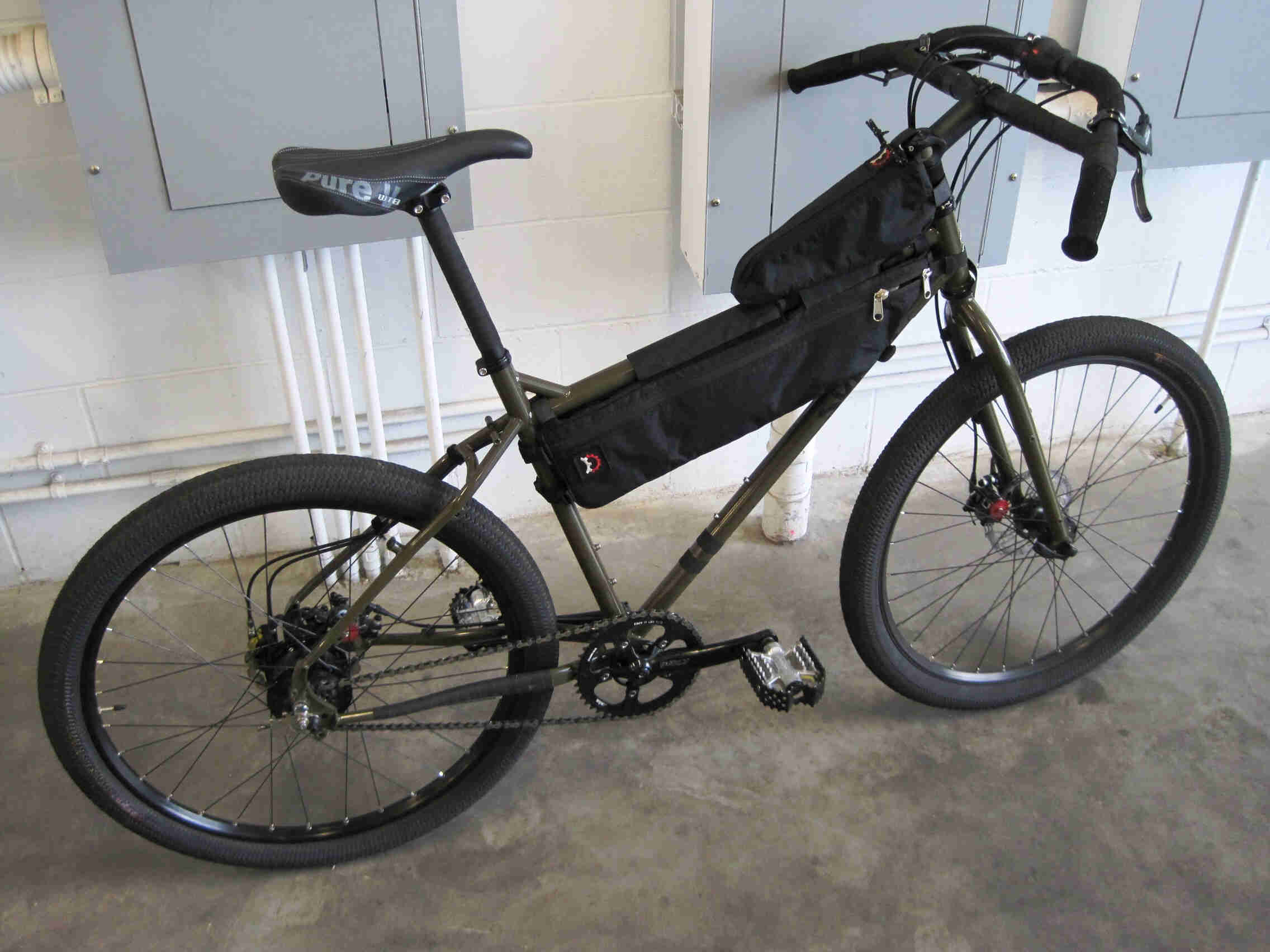 Right side view of a Surly Troll bike with a frame pack, parked on a cement floor, leaning on a wall inside a warehouse